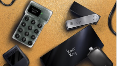 The image shows a flat-lay arrangement of hardware wallet components on a cork surface, including a transparent keypad device displaying "Entropy Bullets," a "Ledger" USB device, a "Keep Key" hardware wallet, a key-shaped item labeled "SEEDSIGNER," and a partial Trezor device. The collection humorously assembles pieces from various crypto security brands into what appears to be a makeshift SeedSigner, fitting the post's joke about an "unintentional ad for @SeedSigner."