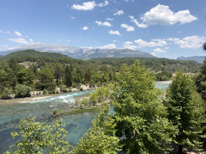 A scenic river flows through a lush green valley with rugged mountains rising in the distance under a bright blue, partly cloudy sky. The image evokes a peaceful, sunny Thursday morning perfect for enjoying nature's beauty.