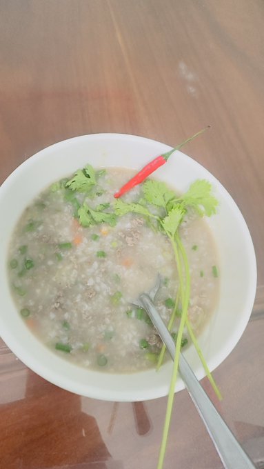 A bowl of Vietnamese-style porridge (cháo) garnished with fresh cilantro, green onions, and a red chili pepper, with a spoon resting in the bowl on a wooden surface.