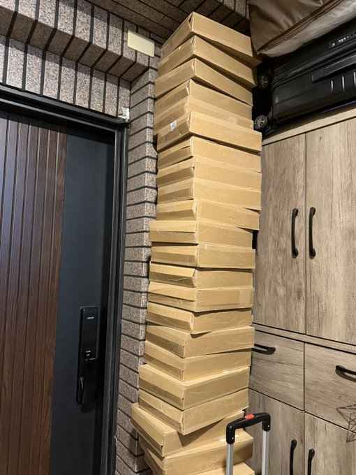 A photograph showing a tall stack of flat, brown cardboard boxes piled up against a brick wall in an entryway, next to a door and wooden cabinets with a suitcase on top.