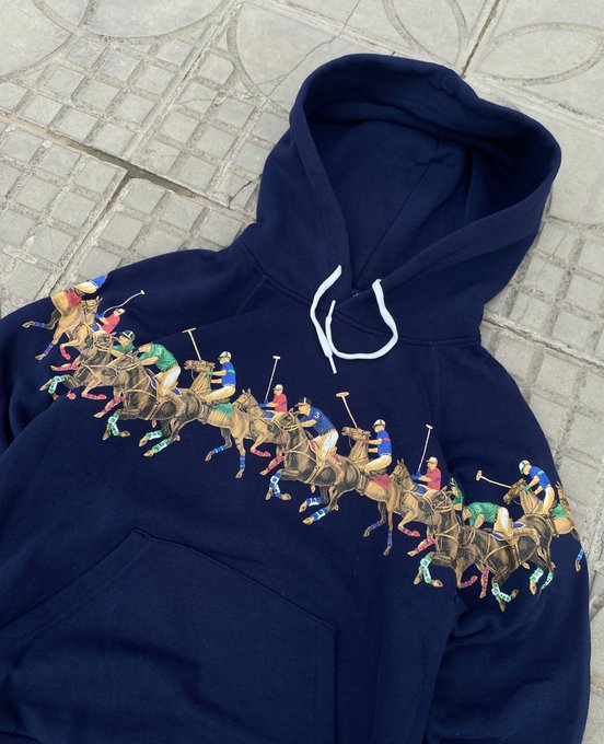 A navy blue hooded sweatshirt lies on a tiled pavement, featuring a vibrant, repeating graphic print of polo players on horseback arranged in a diagonal line across the chest, with white drawstrings visible at the neck.