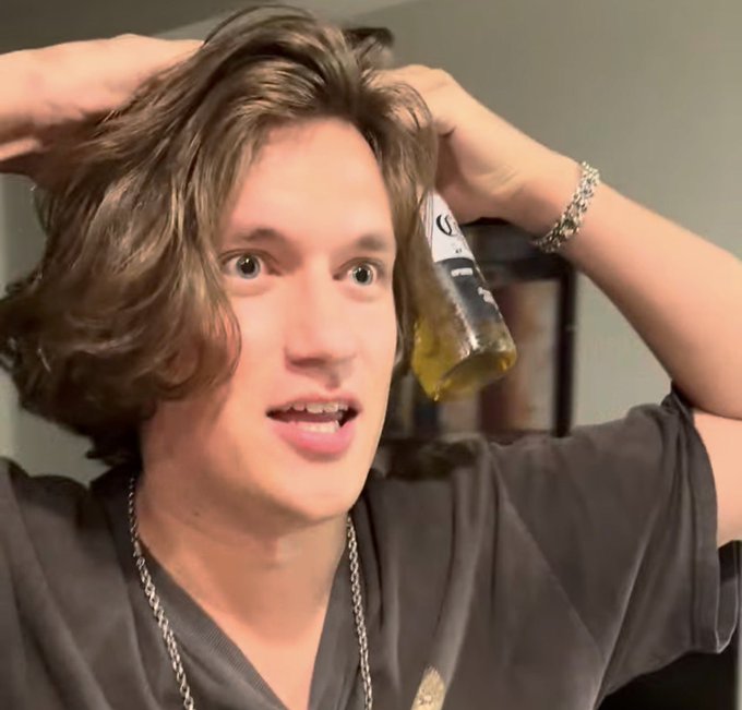 A young man with wavy brown hair holds both hands behind his head in a startled expression while holding a partially empty Corona beer bottle near his face.