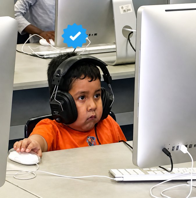 A young boy with headphones using a computer mouse while looking at an iMac monitor in what appears to be a computer lab, with a blue verification checkmark overlaid.