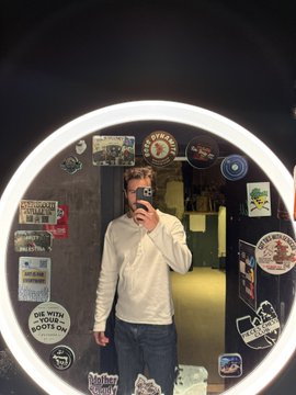 A person taking a mirror selfie in a bathroom covered with various stickers and a round LED light around the mirror.