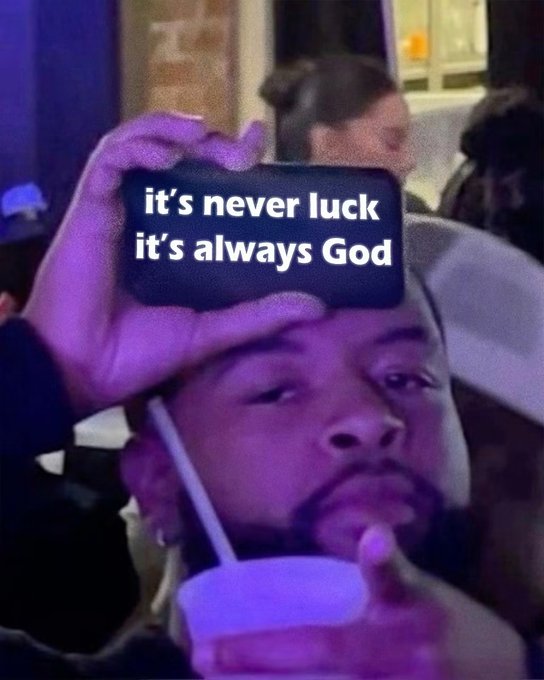 A man in a dimly lit setting sips from a cup while someone holds a phone displaying the caption 'it's never luck it's always God.'