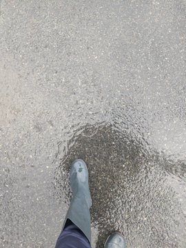 A person wearing dark waterproof boots and navy pants steps through a wet, puddle-covered pavement on a rainy day, splashing water as they walk. The image captures the rainy conditions described in the post, where the user is testing long boots with running shoe insoles for comfort during a wet walk.