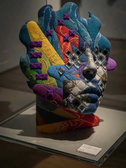 A colorful mixed-media textile sculpture of a face with puzzle piece patterns, metallic silver elements, and mirrored squares is displayed on a glass case with a small label.