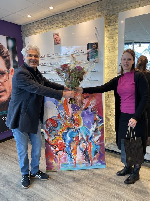 Two people smile as they exchange a bouquet of flowers in front of a vibrant abstract painting at what appears to be an eyewear store, with displays of glasses visible in the background.