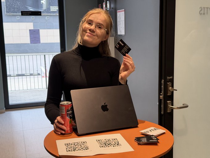 A woman in a black turtleneck poses with a MacBook laptop, holding up a card and a canned drink, with QR codes visible on the table.