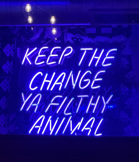 A blue neon sign reading 'KEEP THE CHANGE YA FILTHY ANIMAL' hanging against a patterned wall with string lights.
