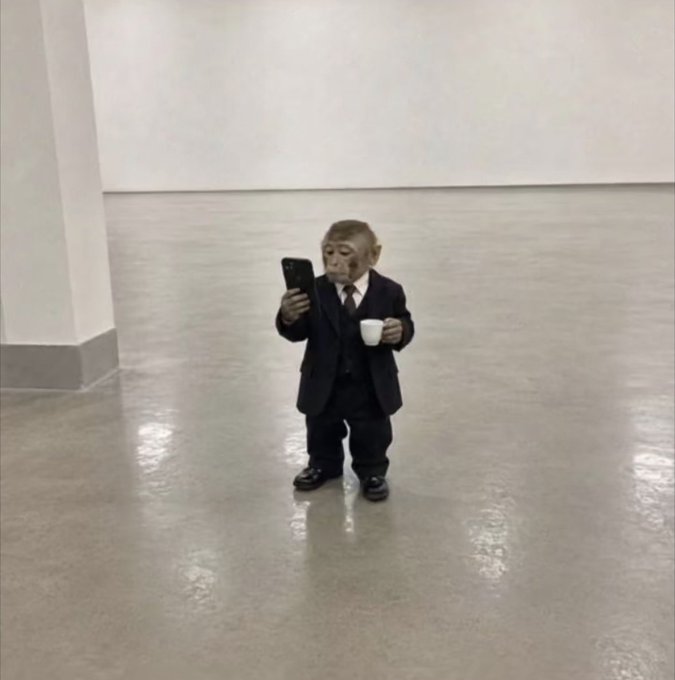 A small monkey dressed in a formal black suit and tie stands alone in a minimalist white gallery space, holding a smartphone in one hand and a white cup in the other, whimsically juxtaposing corporate attire with a playful "businessman" persona that matches the post's humorous crypto-focused tone about BeldexCoin.