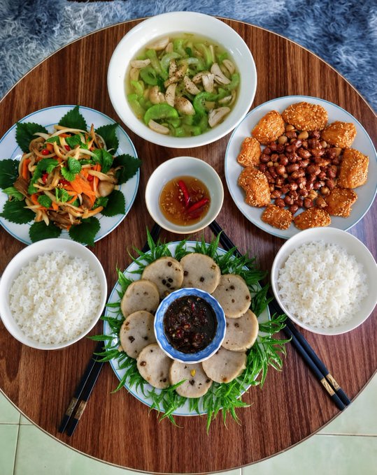 An overhead view of a Vietnamese meal on a round wooden table, featuring two bowls of white rice, a dish of sliced meat with chili dipping sauce, a vegetable soup, a plate of peanuts with tofu crackers, and a fresh herb salad.