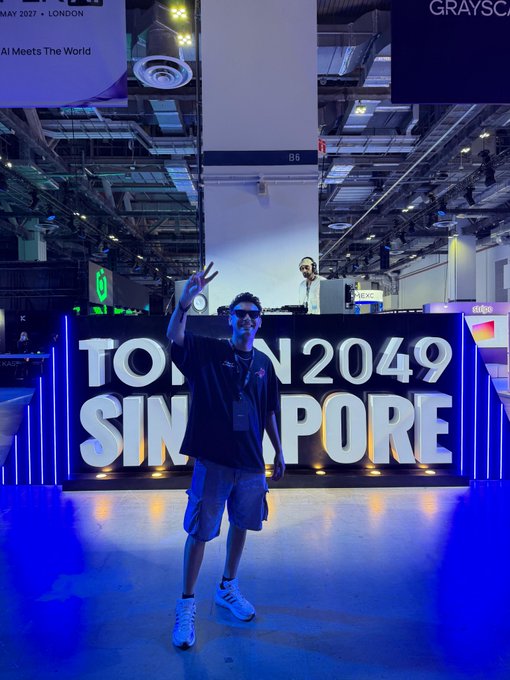 A man in a black shirt, blue shorts, and sunglasses posing with a peace sign in front of a large illuminated 'TOKEN2049 SINGAPORE' sign at a conference venue.