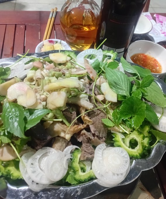 A large silver platter holds a Vietnamese dish with beef, bitter melon, lotus root slices, herbs, and peanuts, accompanied by a small bowl of red dipping sauce and a glass of iced tea.