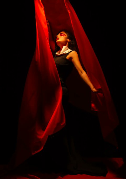 A dramatic, low-key photograph of a woman in a black sleeveless dress surrounded by flowing red fabric, with red lighting illuminating her face and arms against a black background.