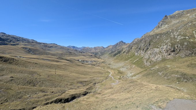 A sweeping mountain landscape featuring rolling grassy hills and rocky peaks under a clear blue sky, with a winding road traversing the valley below and a contrail visible in the sky.