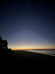 A serene night beach scene with a starry sky, faint silhouettes of trees on the left, and distant city lights glowing along the horizon over calm water.