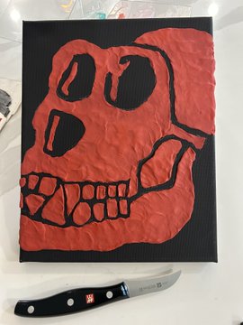 A red and black painted artwork depicting a stylized skull with teeth, displayed on a black background next to a small knife.