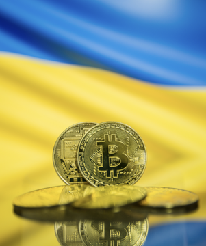 Two physical Bitcoin tokens sit on a reflective surface against a waving Ukrainian flag, visually linking the $8.3 million in seized USDT transferred to Ukraine's state management with the country's emerging stance on cryptocurrency regulation.