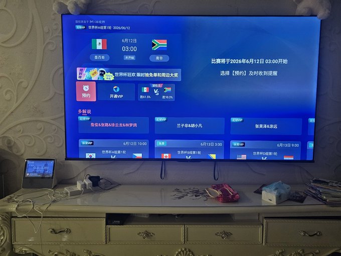 The image shows a flat screen TV mounted on top of a white dresser, displaying a foreign language channel with various countries' flags displayed. The TV is turned on, and there are several channels available for viewing. A remote control can be seen in front of the television, suggesting that it's ready to be used. 

In addition to the TV, there are some books placed nearby, indicating that this might be a living room or entertainment area where people gather to watch TV shows or movies. The presence of multiple channels and the remote control suggests that the viewer has various options for choosing what they want to watch.
