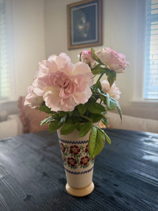 A bouquet of pink roses sits in a decorative ceramic vase with red floral patterns, placed on a dark wooden table in a room with a framed picture on the wall.