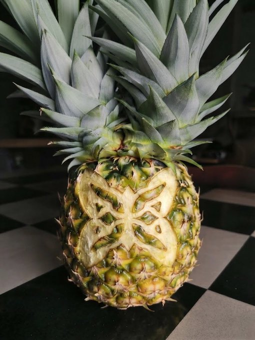 A photograph of a carved pineapple with a geometric design cut into its side, sitting on a black and white checkered surface.