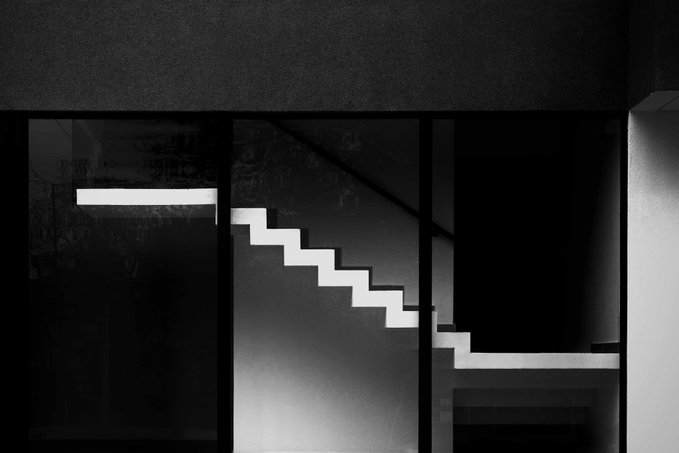 A stark black-and-white photograph captures a modern staircase illuminated by dramatic lighting, viewed through a glass partition, symbolizing the metaphorical journey of life mentioned in the post—each illuminated step representing progress along the human path.