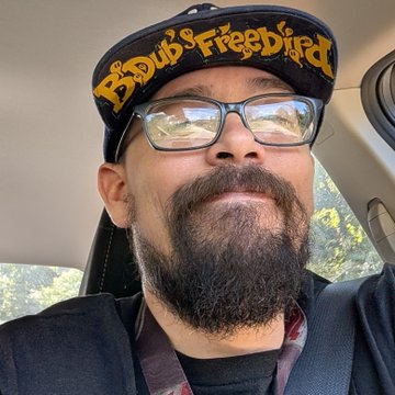 A bearded man wearing glasses and a "B's Dubs & Freebird" cap smiles from inside a car, capturing his excitement as he heads to the airport for his journey to Salt Lake City and LA for "Solana Summer House."