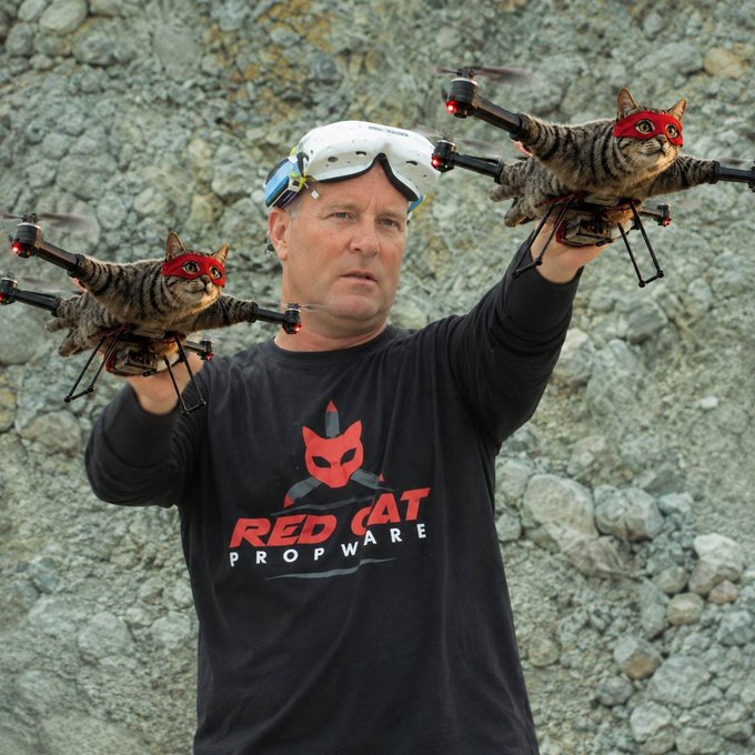 A man wearing a 'Red Cat Propware' shirt and FPV goggles holds two small drones, each with a tabby cat wearing a red mask attached to it, against a rocky background.