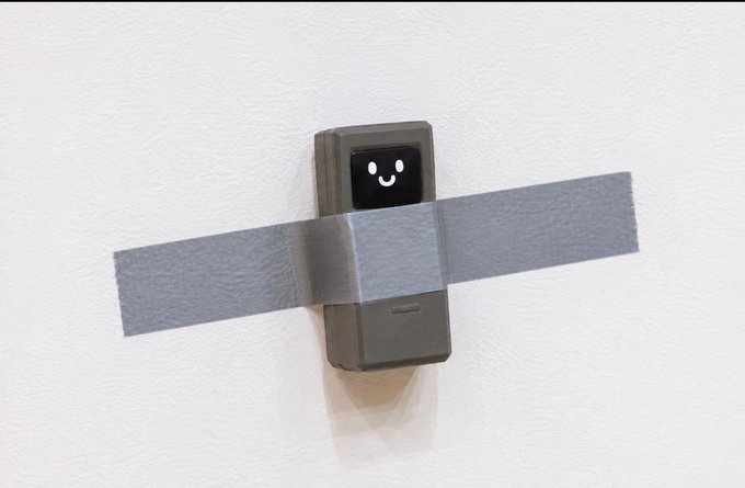A small electronic device (appearing to be a Ring doorbell) mounted on a white wall with a smiley face displayed on its screen, taped across with silver duct tape.