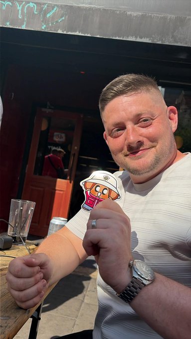 A man smiles at the camera while holding up a cartoon chimp sticker wearing a sailor hat, appearing to be outside a venue in Manchester. A partially visible sign in the background reads "YEAR," and the relaxed outdoor setting matches the post's description of a sunny "gm" meetup with the Chimpers x Fugz communities.