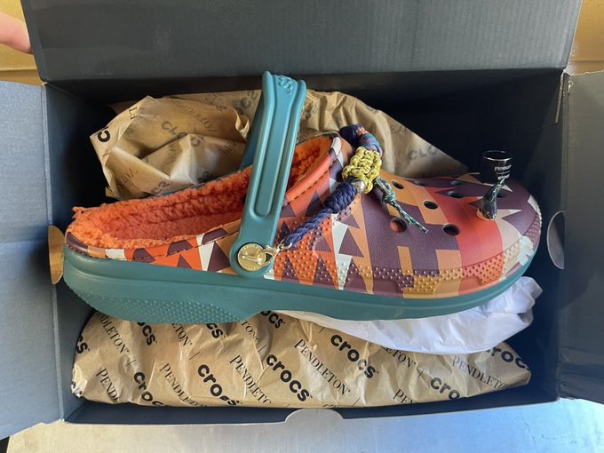 A Pendleton Crocs shoe with a colorful patterned upper, teal heel strap, and colorful laces, shown in its branded shoe box with tissue paper.