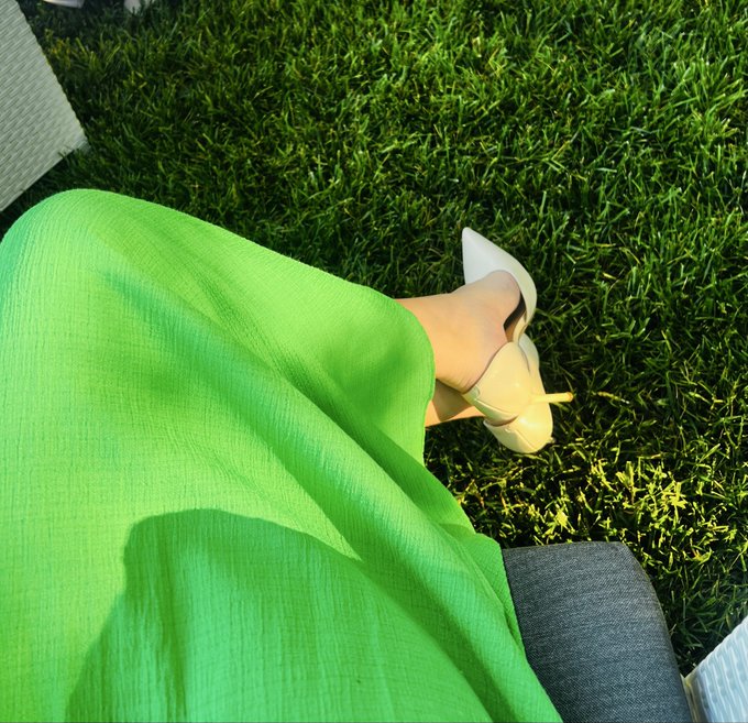 A downward view of a person wearing a bright green dress and white/nude heels sitting on grass with sunlight casting shadows on their lap.