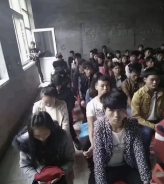 A crowded classroom filled with students seated closely together, with some standing near the windows in a dimly lit room.