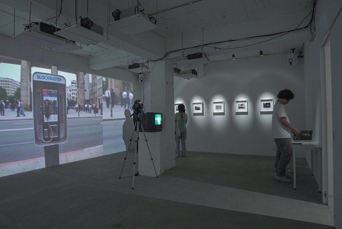 An indoor art gallery displays framed photographs on a white wall, a large street scene projection with a 'BLOCKBUSTER' phone booth, and two visitors viewing the exhibits.