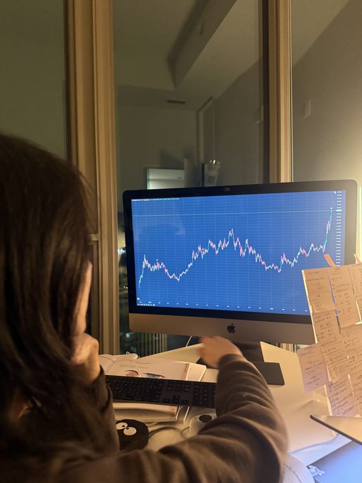 The image shows a woman sitting at a desk with a monitor displaying stock market graphs. The stock prices are changing, indicating that she might be a trader or interested in financial markets. In addition to the monitor, there's also an external keyboard visible on the table. The presence of the woman and her focus on the computer screen suggests that she is actively engaged in analyzing the stock data and making decisions based on the fluctuations displayed on the monitor.