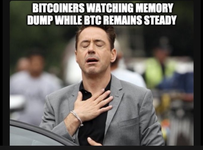 Robert Downey Jr. clutches his chest in a meme captioned "BITCOINERS WATCHING MEMORY DUMP WHILE BTC REMAINS STEADY," humorously capturing the post's sentiment of exasperated relief as $BTC holders endure unrelated market chaos.