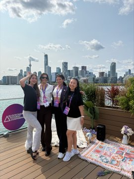 A group of four women wearing lanyards posing for a photo on a wooden deck with a city skyline in the background; a green sign reading 'ADVENTURE AWAITS' is visible on the left.