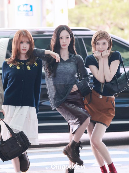 Three women pose together on a street with a black car behind them; one wears a navy cardigan, the middle wears a gray top with a brown skirt, and the right wears a black top with a brown skirt, with an 'Omgoddess' watermark at the bottom.