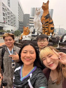 A selfie of four smiling people posing in front of cat statues and city buildings, likely in a public square or park.