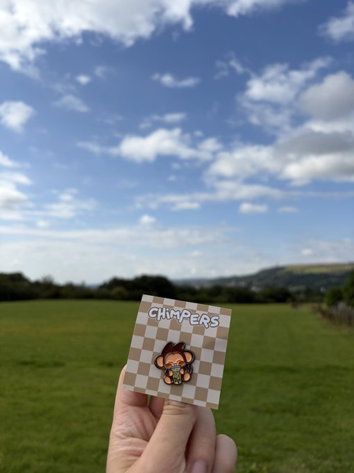 A hand holds up a small card with a brown and beige checkerboard pattern featuring a cartoon character and the text "CHIMPERS" against a backdrop of a green field and blue cloudy sky.