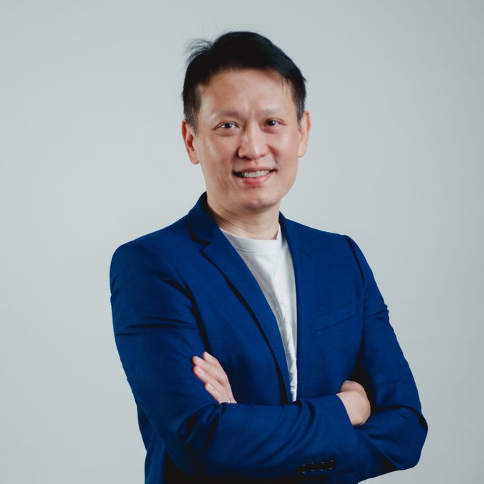 A confident man with short black hair, wearing a blue blazer over a white t-shirt, stands with arms crossed against a light gray background, smiling at the camera. The image appears to be a professional portrait of Richard Teng, the Binance co-CEO referenced in the accompanying post about the CLARITY Act and regulatory concerns in the crypto industry.