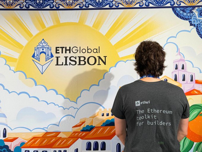 A person viewed from behind wearing a gray t-shirt reading 'ethui - The Ethereum toolkit for builders' while standing in front of the colorful ETHGlobal LISBON event backdrop.