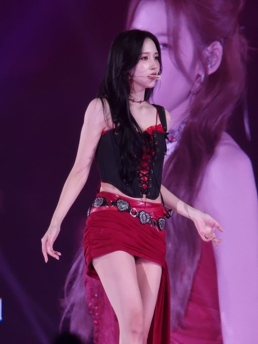 A performer with long dark hair, wearing a black corset top, red skirt, and ornate belt, sings into a headset microphone on stage under pink-purple lighting, fitting the "Mina vampire propaganda" aesthetic with her gothic, blood-red ensemble evoking a vampiric vibe.