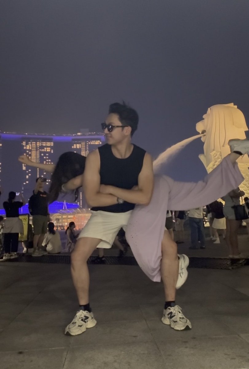 A man wearing a black tank top, white shorts, and sunglasses poses with his arms crossed while holding a woman in a lavender dress across his lap, with the Merlion statue and Marina Bay Sands visible at night in the background.