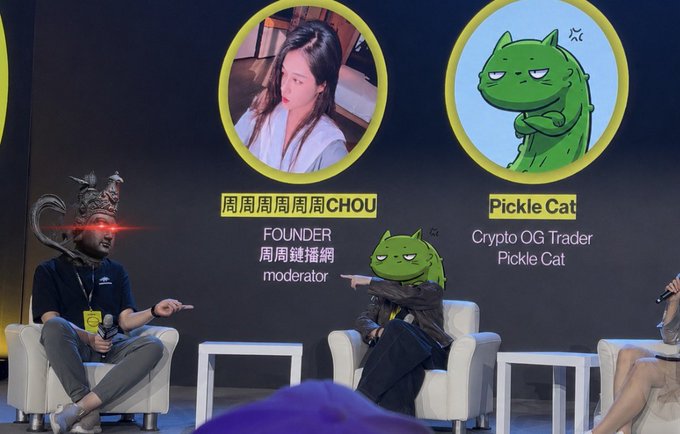 Two masked figures sit on stage at a crypto panel discussion, with a screen behind them displaying profiles for "周周周周周周CHOU" (Founder, moderator) and "Pickle Cat" (Crypto OG Trader), matching the humorous moment from the post where the host asks who's making money and the two speakers point at each other.