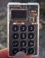 A close-up of a small DIY electronic device labeled 'Packet Monitor,' featuring a digital display screen and a numeric keypad held between fingers.