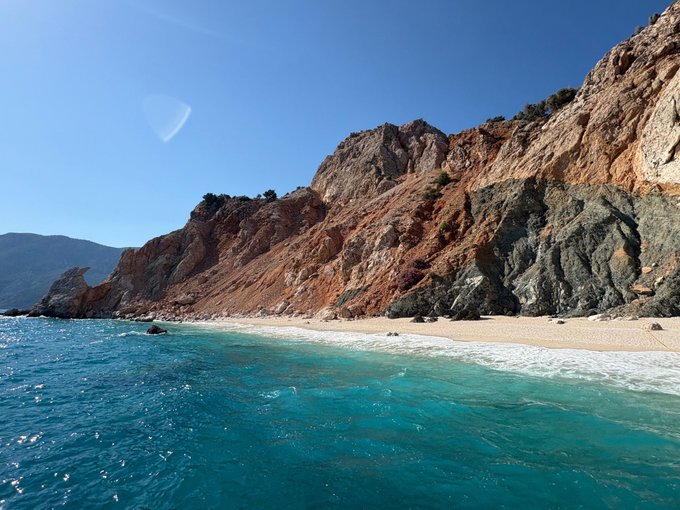 A picturesque coastal scene features turquoise waters lapping against a secluded sandy beach nestled beneath dramatic, rust-colored rocky cliffs under a clear blue sky, capturing the natural beauty of a remote shoreline.