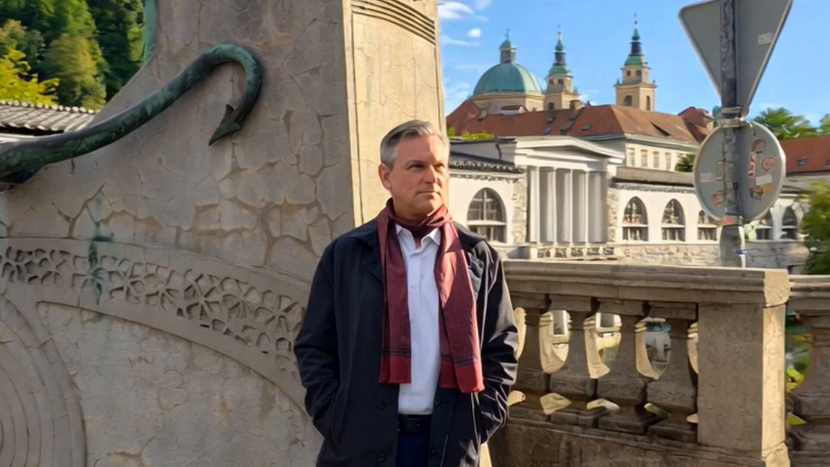 The image features a man standing next to a stone building with a large archway. The man appears to be wearing a red scarf, black coat, and white shirt. He seems to be looking off into the distance or observing something happening nearby. The background of the image shows a cityscape with other buildings in the vicinity.