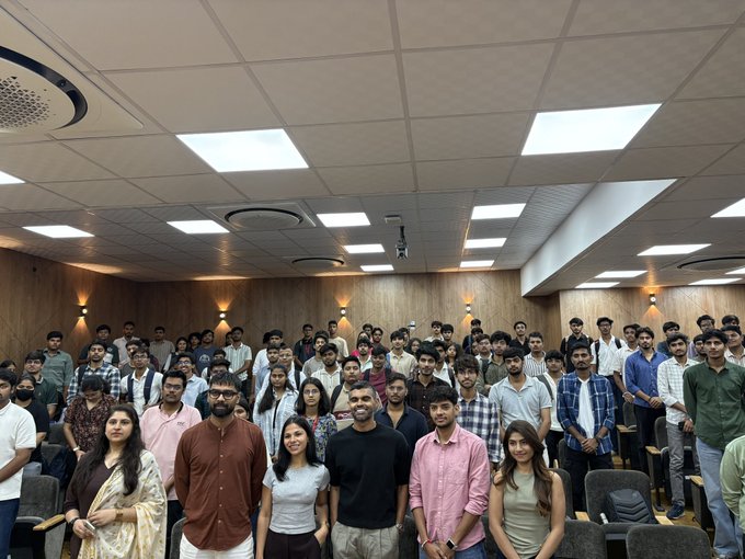 A large group of students and professors pose for a group photo inside a brightly lit auditorium or lecture hall with wood-paneled walls and rows of seats.